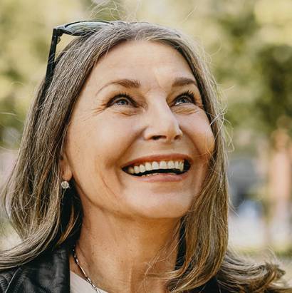 Cheerful overjoyed middle aged woman wearing sunglasses on her head and black leather jacket over white blouse enjoying peaceful beautiful morning while waking in park, looking up with broad smile