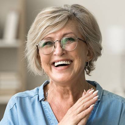 Cheerful pretty older woman in elegant glasses sitting on cozy home couch, smiling with perfect white teeth, laughing with hands at chest gesture, enjoying leisure, comfort, having fun
